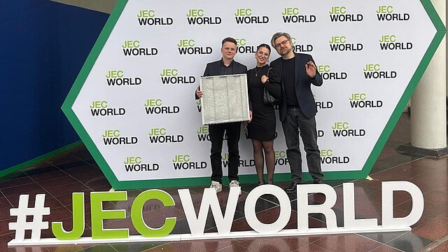 trzy osoby ubrane na galowo stoją obok siebie, jedna osoba trzyma materiał